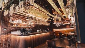 Un bar con pared de ladrillo, mostrador de madera y flores blancas colgantes. Se observan estantes con botellas y un área de asientos con sillas y una mesa.