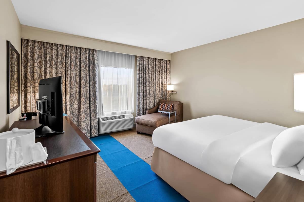 Habitación de hotel con cama, escritorio con televisor, silla y ventana con cortinas.