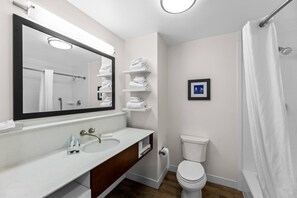 A modern bathroom with a large mirror, white sink, and toilet. There are shelves with folded towels and a framed picture on the wall.