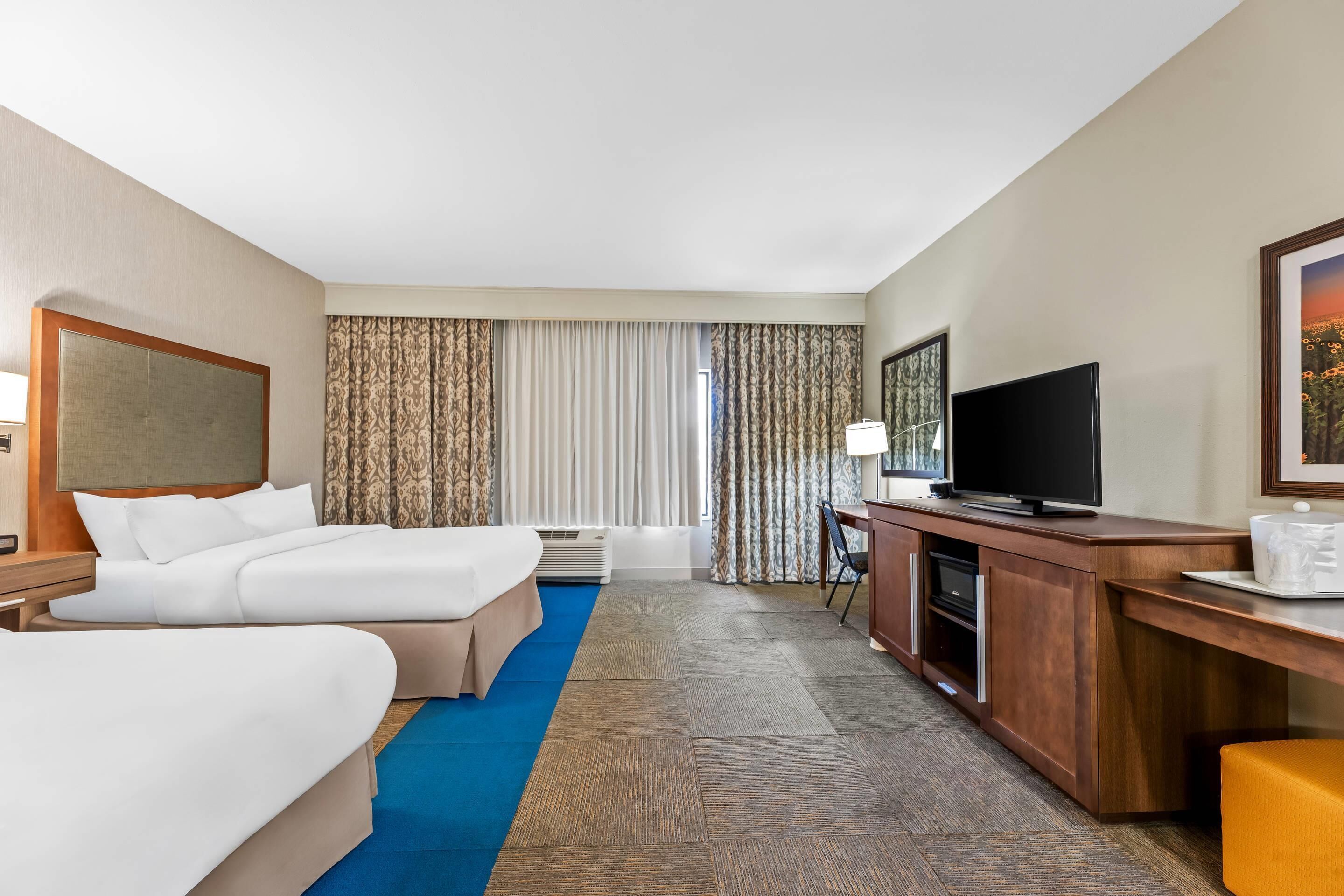 A hotel room with two beds, a desk with a television, and a large window with curtains.