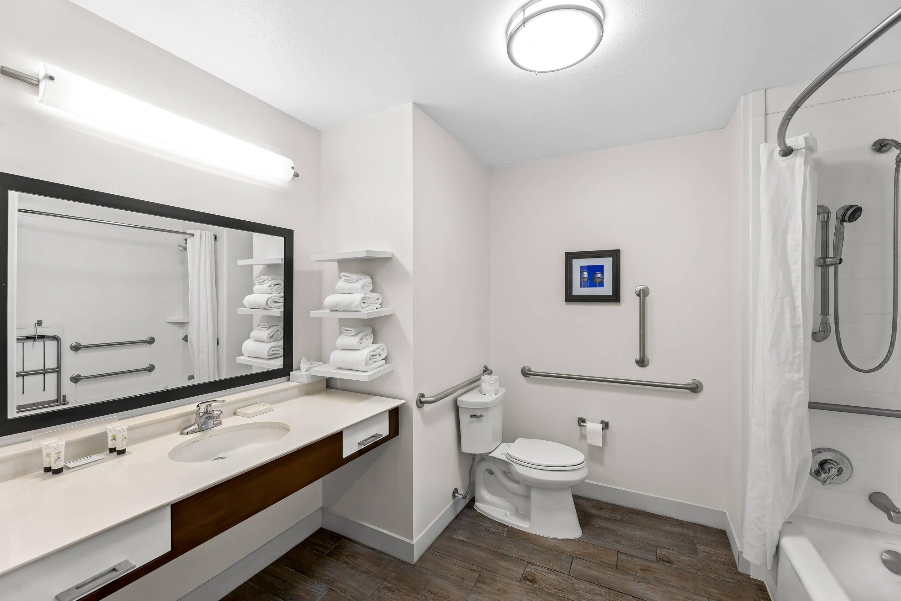 A modern bathroom with a large mirror, white fixtures, a shower with grab bars, and a toilet with a handrail.