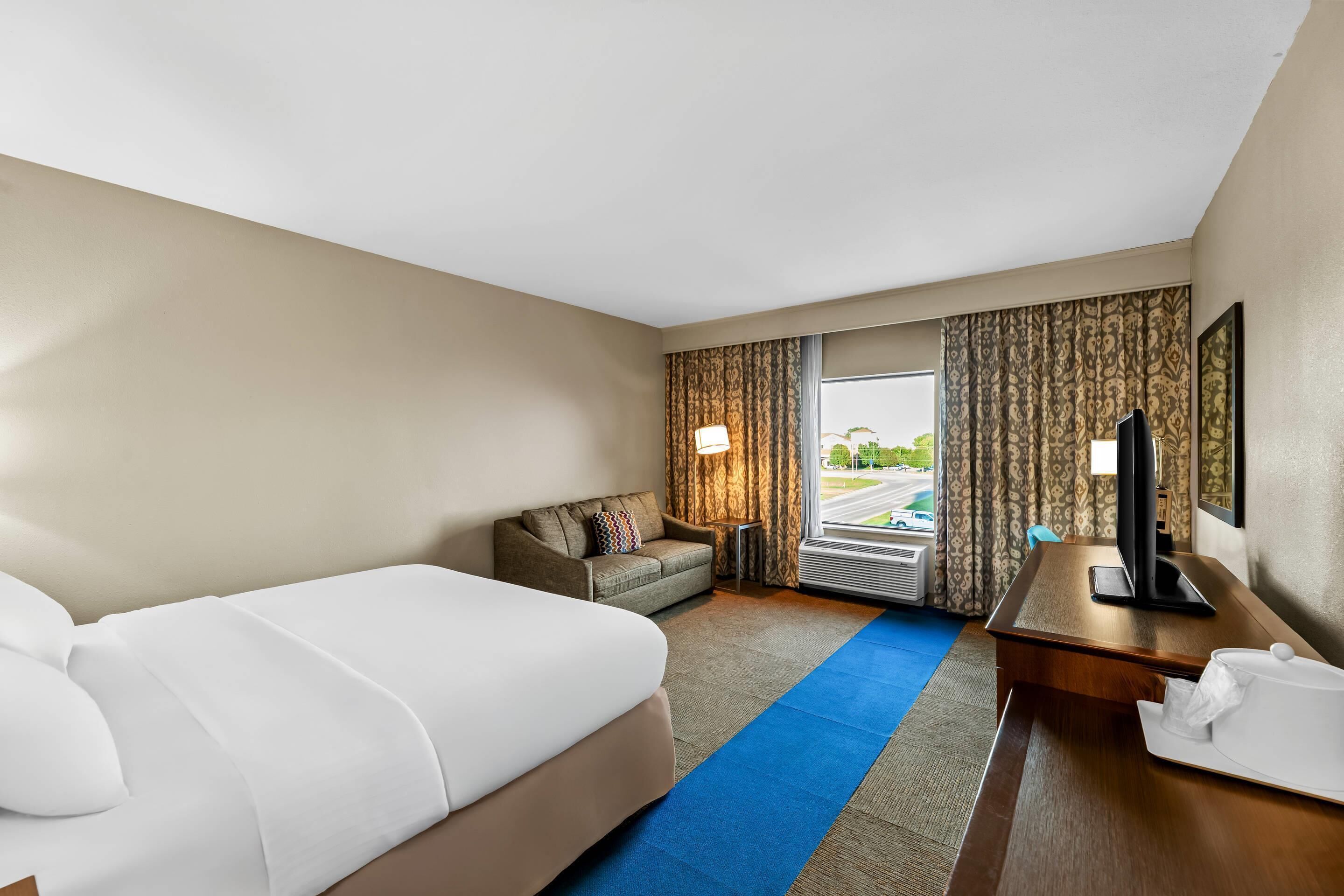 A hotel room with a large bed, a sofa, a desk with a television, and a window with curtains.