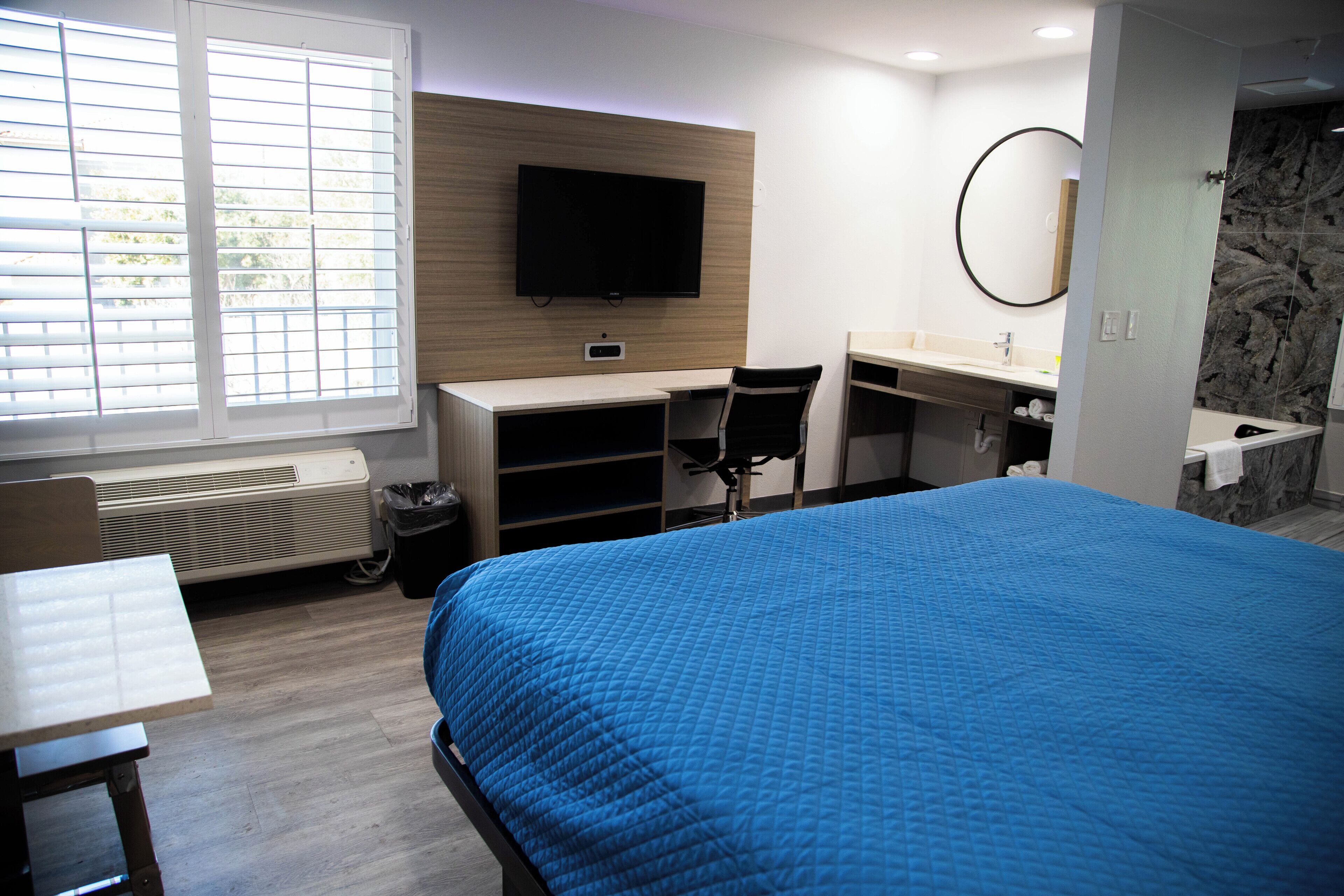 A hotel room with a bed, a desk, a chair, a TV, a bathroom, and a window with blinds.