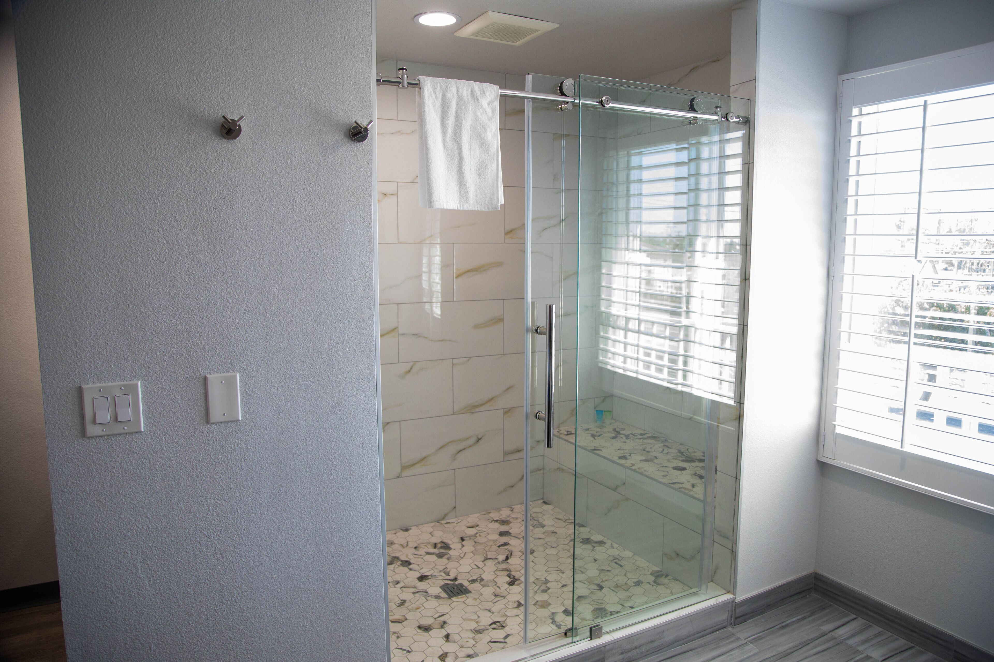 A modern bathroom with a glass-enclosed shower, marble tiles, and a window with blinds.