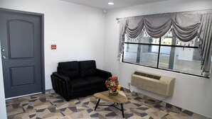 Ein Zimmer mit geometrischem Fußbodenmuster, einer schwarzen Couch, einem kleinen Holztisch mit einer Vase, einem Fenster mit Vorhängen und einer wandmontierten Klimaanlage.