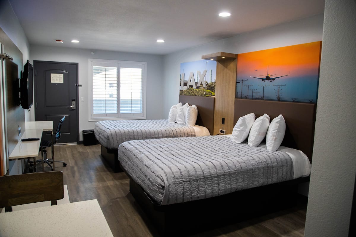 A hotel room with two beds, a desk, and a chair. There is a window with blinds, a door, and a wall-mounted TV.