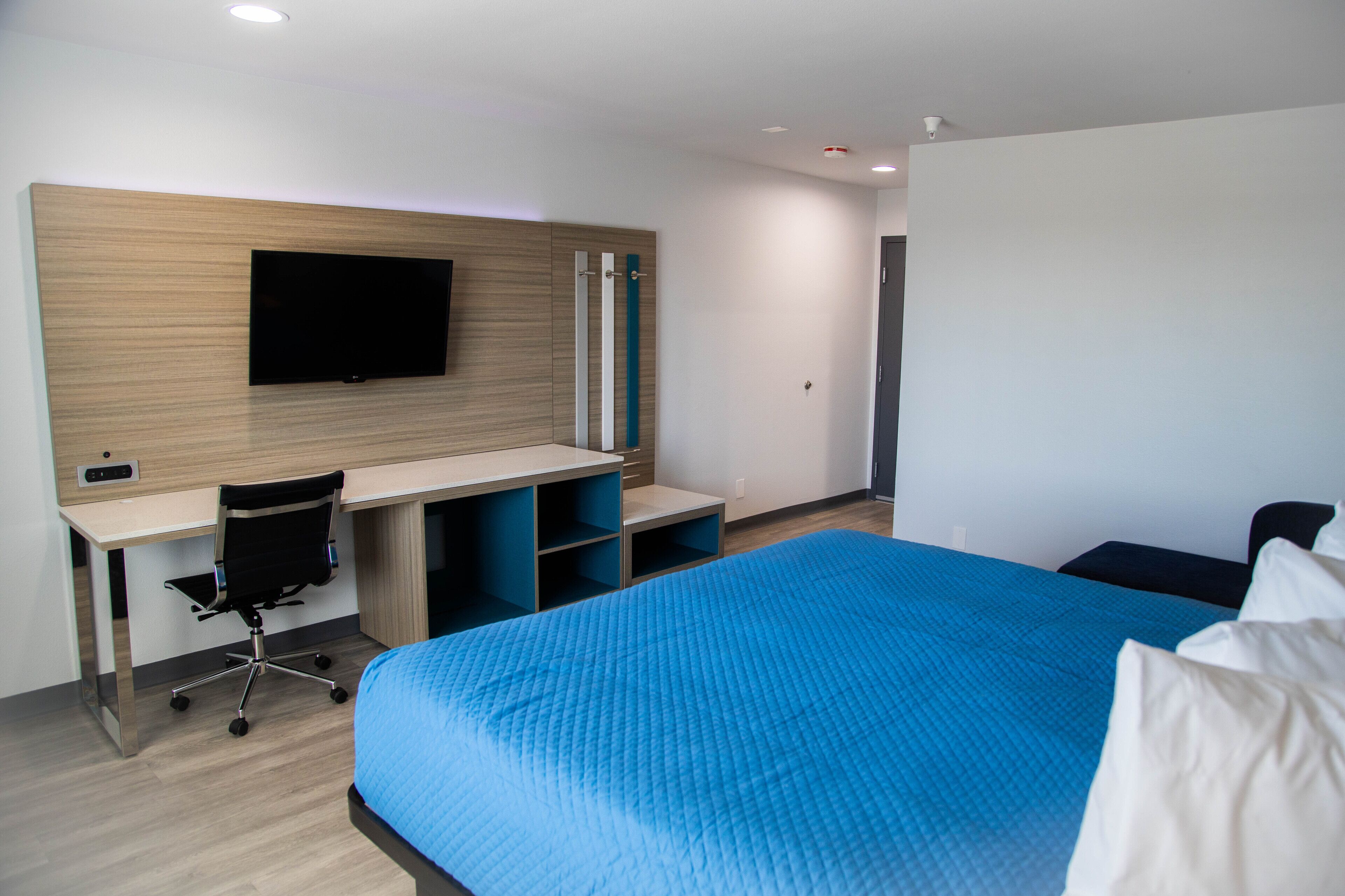 A modern hotel room with a large bed, a desk with a chair, a flat-screen TV mounted on the wall, and a wardrobe.