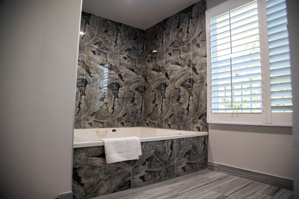 A modern bathroom with a marble wall, a white bathtub, and a window with blinds.