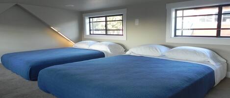 A room with two beds, each with blue bedding and white pillows, a sloped ceiling, and windows with blinds.