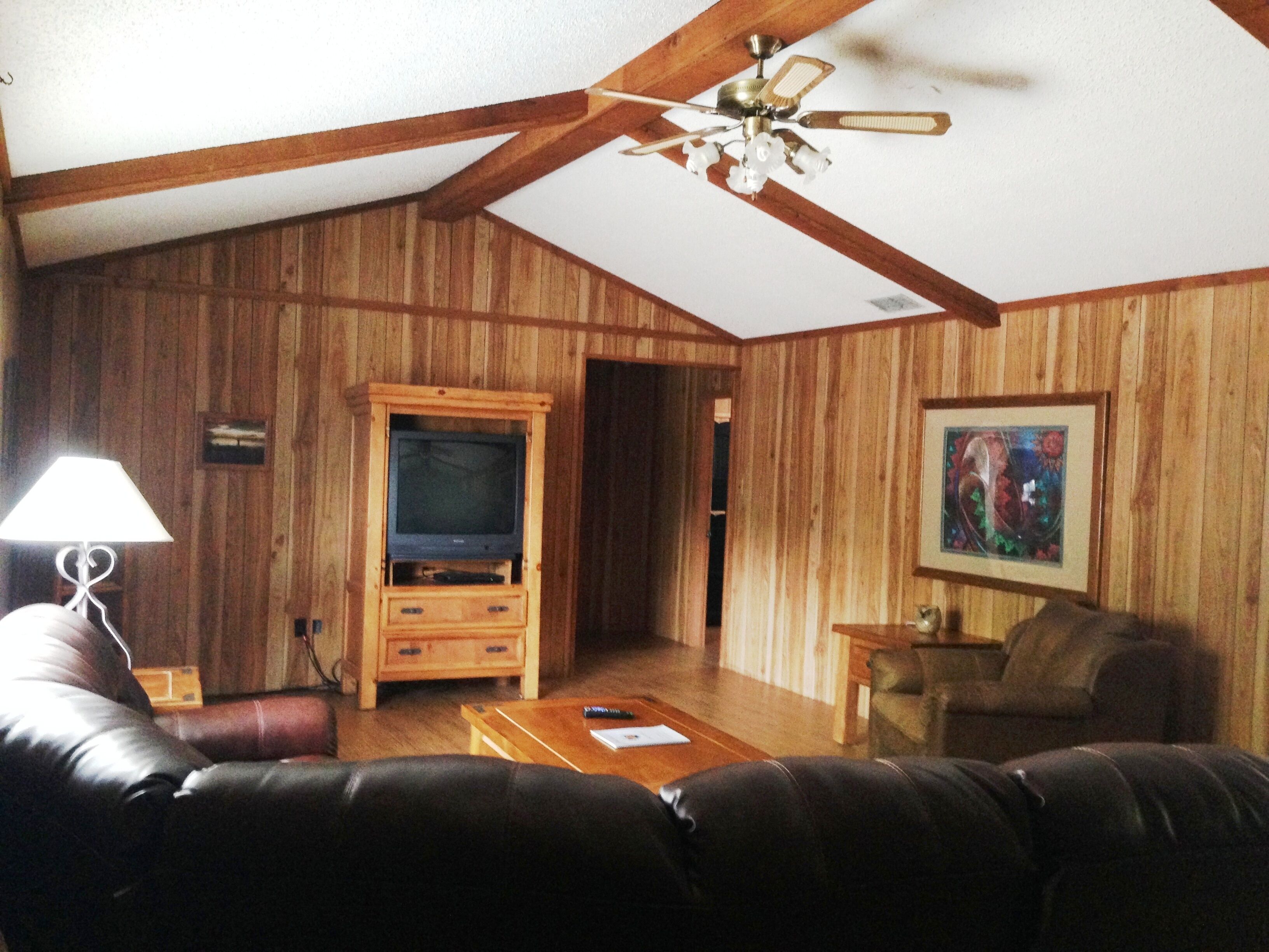 A living room with wooden paneling, a ceiling fan, a television on a wooden cabinet, a coffee table, a sofa, and a painting on the wall.