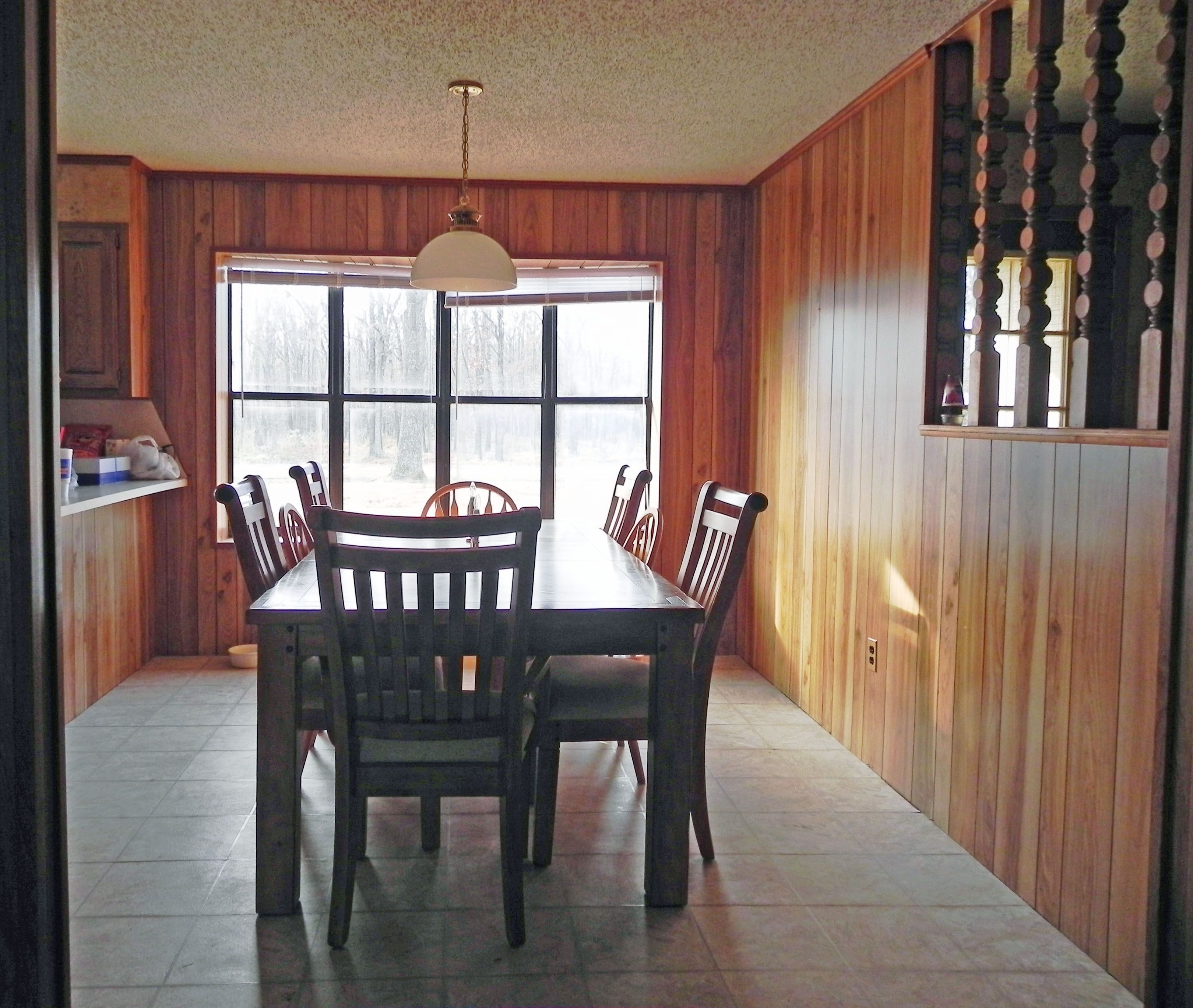 Un comedor con una mesa y sillas de madera, una lámpara colgante y una ventana con vista a árboles.