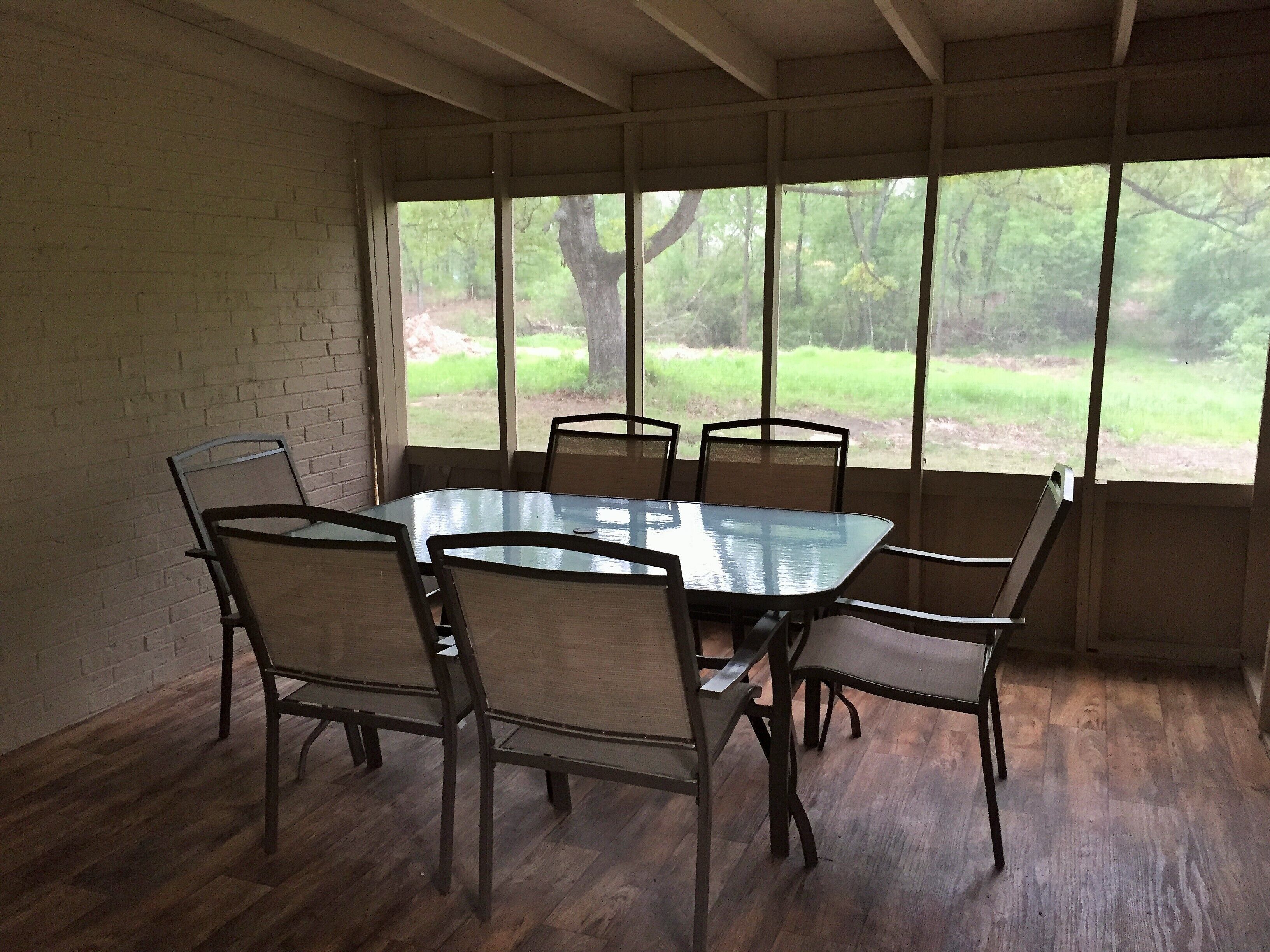 A dining area with a glass table and six chairs, large windows with a view of greenery, and wooden flooring.