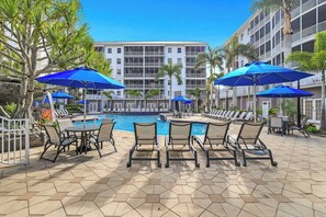 A poolside area with lounge chairs and umbrellas, a pool, and a multi-story building with balconies.