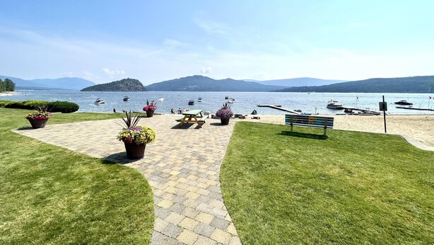 A paved walkway leading to a lakeside area with picnic tables and benches, surrounded by well-maintained grass and potted plants.