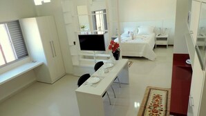 A modern, well-lit kitchen with white cabinetry, a dining area with a table set for two, and a bedroom with a bed and bedside tables. - Hotel Pequeno Mundo II (Campo Verde)