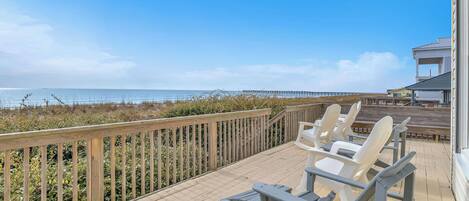 A wooden deck with chairs and tables overlooking a beach and a pier.
