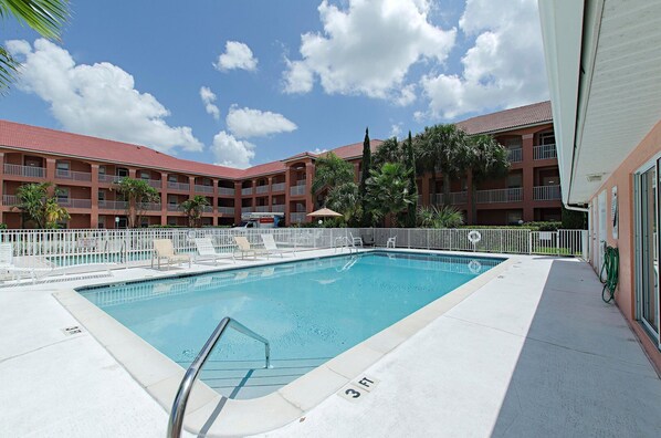 A swimming pool with lounge chairs and a fenced area, surrounded by a multi-story building with balconies.
