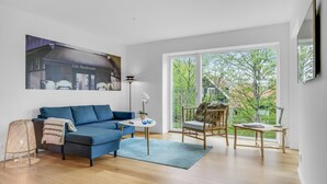 A modern living room with a blue sofa, a small round table, and a chair. There is a large window with a view of trees and a balcony. - Nice apartment in Hornbæk with WiFi (Hornbæk)