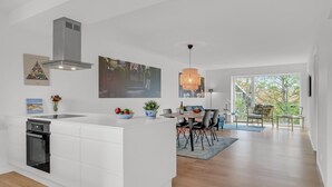 A modern kitchen with white cabinetry, a built-in oven, and a large island with a bowl of fruit. There is a dining area with a table and chairs, and a large window offering a view of the outdoors. - Nice apartment in Hornbæk with WiFi (Hornbæk)