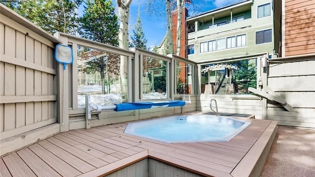 A hot tub with a wooden deck and a glass enclosure, surrounded by trees and a building in the background. - Prime Location Incredible Amenities Hot Tub Wifi (Breckenridge)