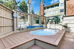 Ein Whirlpool mit Holzterrasse und Glasumrandung, umgeben von Bäumen und einem Gebäude im Hintergrund.
