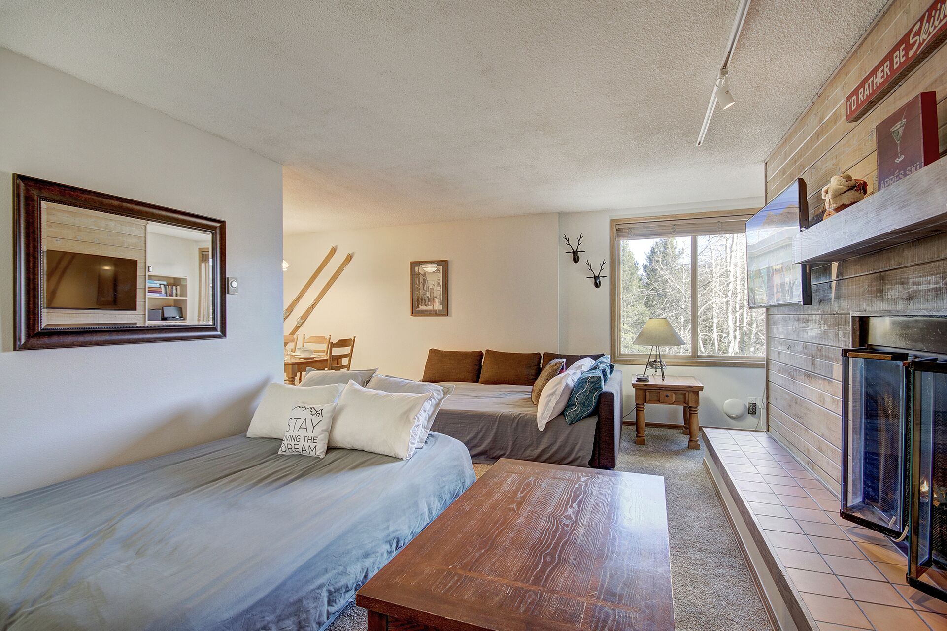 A cozy room with a bed, a wooden table, a fireplace, and a window with a view of trees.