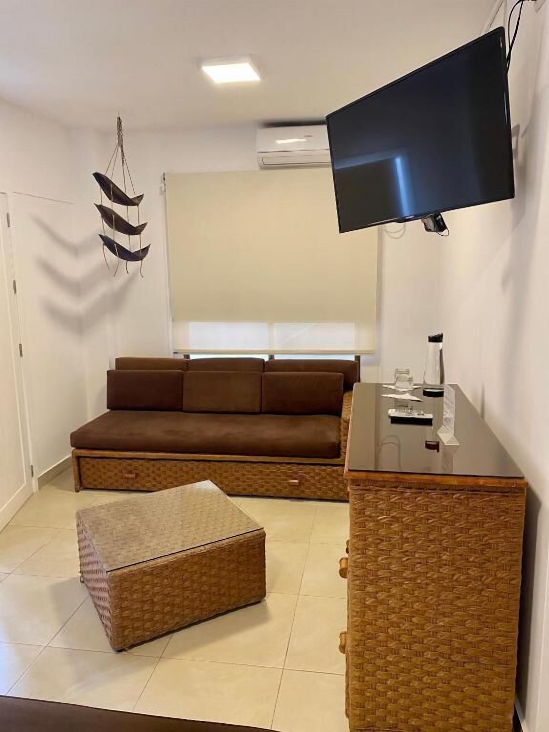 A living room with a brown sofa, a wicker ottoman, a flat-screen TV mounted on the wall, and a small table with a lamp.