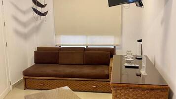 A living room with a brown sofa, a wicker ottoman, a flat-screen TV mounted on the wall, and a small table with a lamp.