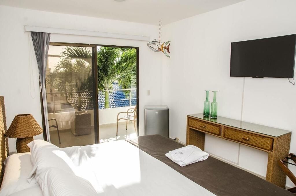 Habitación de hotel con cama, escritorio, silla, televisor y vistas a palmeras a través de una puerta corrediza de vidrio.