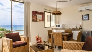Una habitación con mobiliario de mimbre, comedor, cocina y un amplio ventanal con vista al mar.