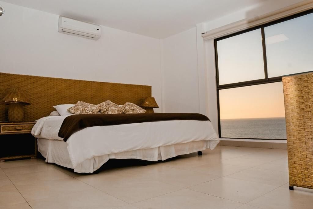 A bedroom with a large window, a bed with white bedding, a nightstand with a lamp, and a woven headboard.