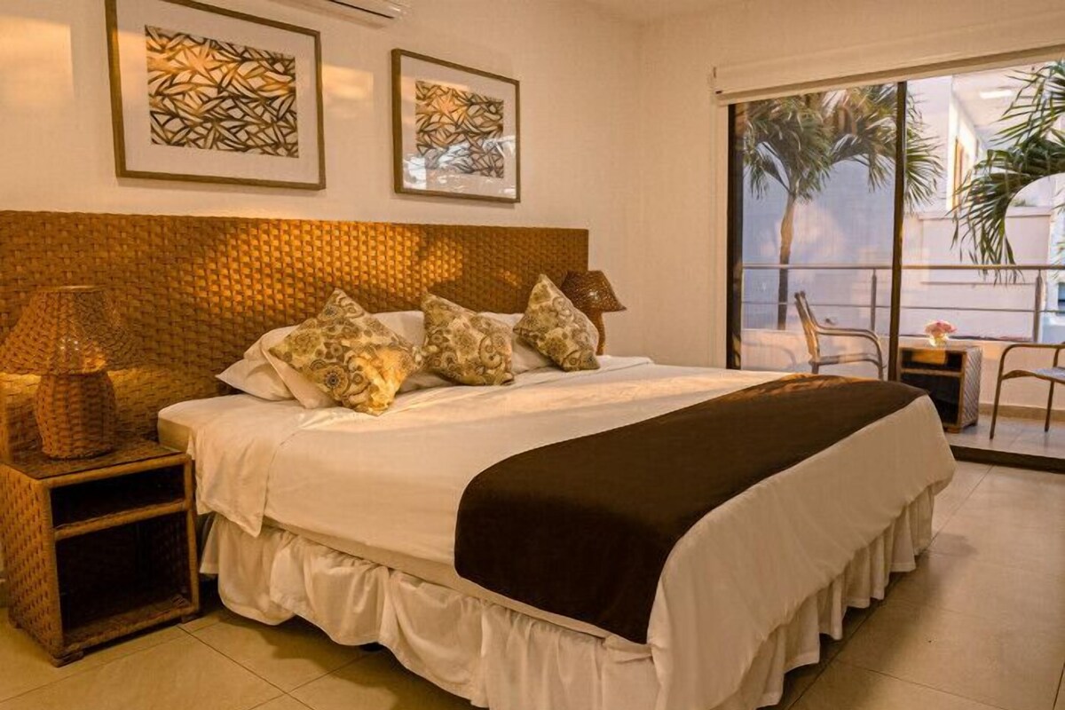 Habitación de hotel con una cama grande, una mesita de noche, una lámpara y dos cuadros en la pared.