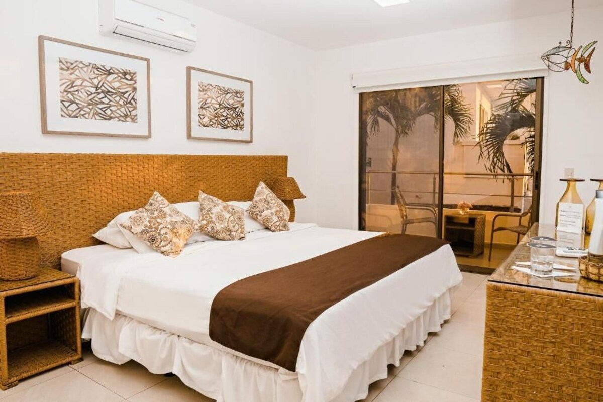 Habitación de hotel con una cama grande, una mesita auxiliar pequeña, una silla de mimbre y vistas a palmeras a través de una puerta corredera.
