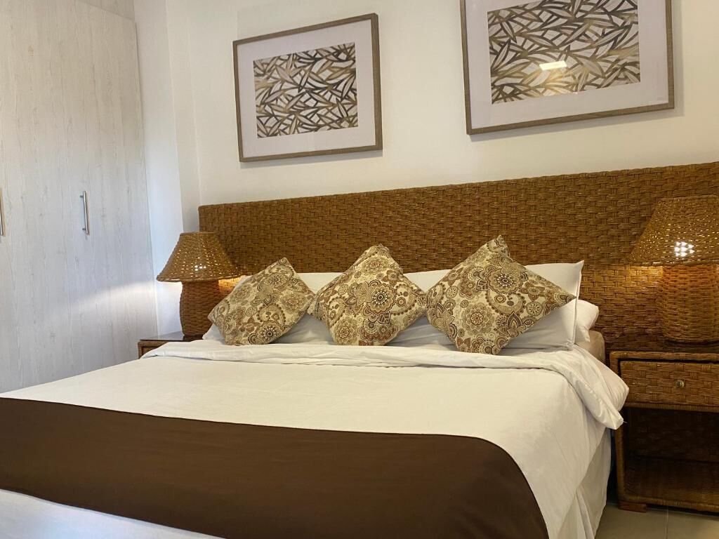 Cama bem arrumada com almofadas estampadas, cabeceira marrom e duas luminárias fixadas na parede.