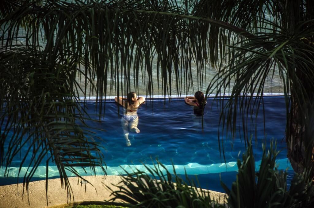 A swimming pool with two people in the water, surrounded by palm trees.
