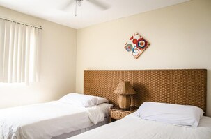A hotel room with two single beds, a woven headboard, a woven nightstand, a lamp, and a wall art piece.