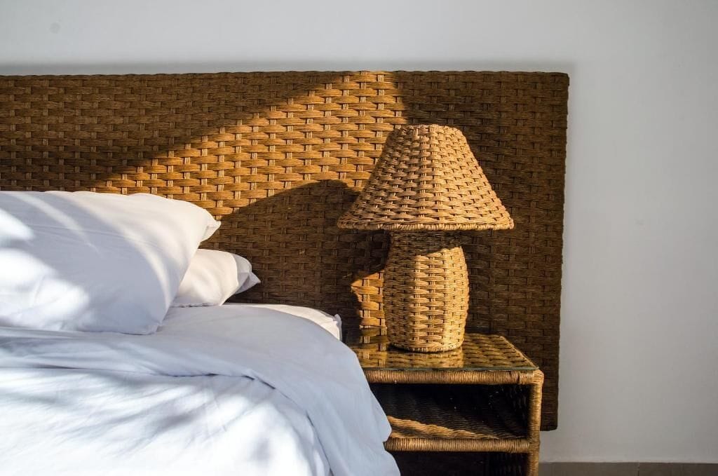 Ein Bett mit weißer Bettwäsche, ein Korbnachtisch mit einer gewobenen Lampe und ein gewobener Wandvorhang als Kopfteil.