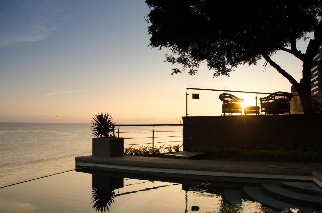 A sunset view from a balcony overlooking the sea, with a pool and outdoor seating area.
