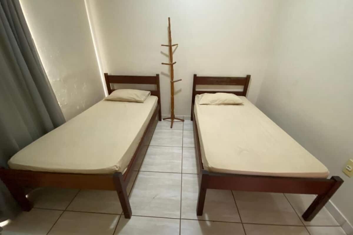 Two single beds with wooden frames and beige mattresses in a small room with tiled flooring and a window with curtains.