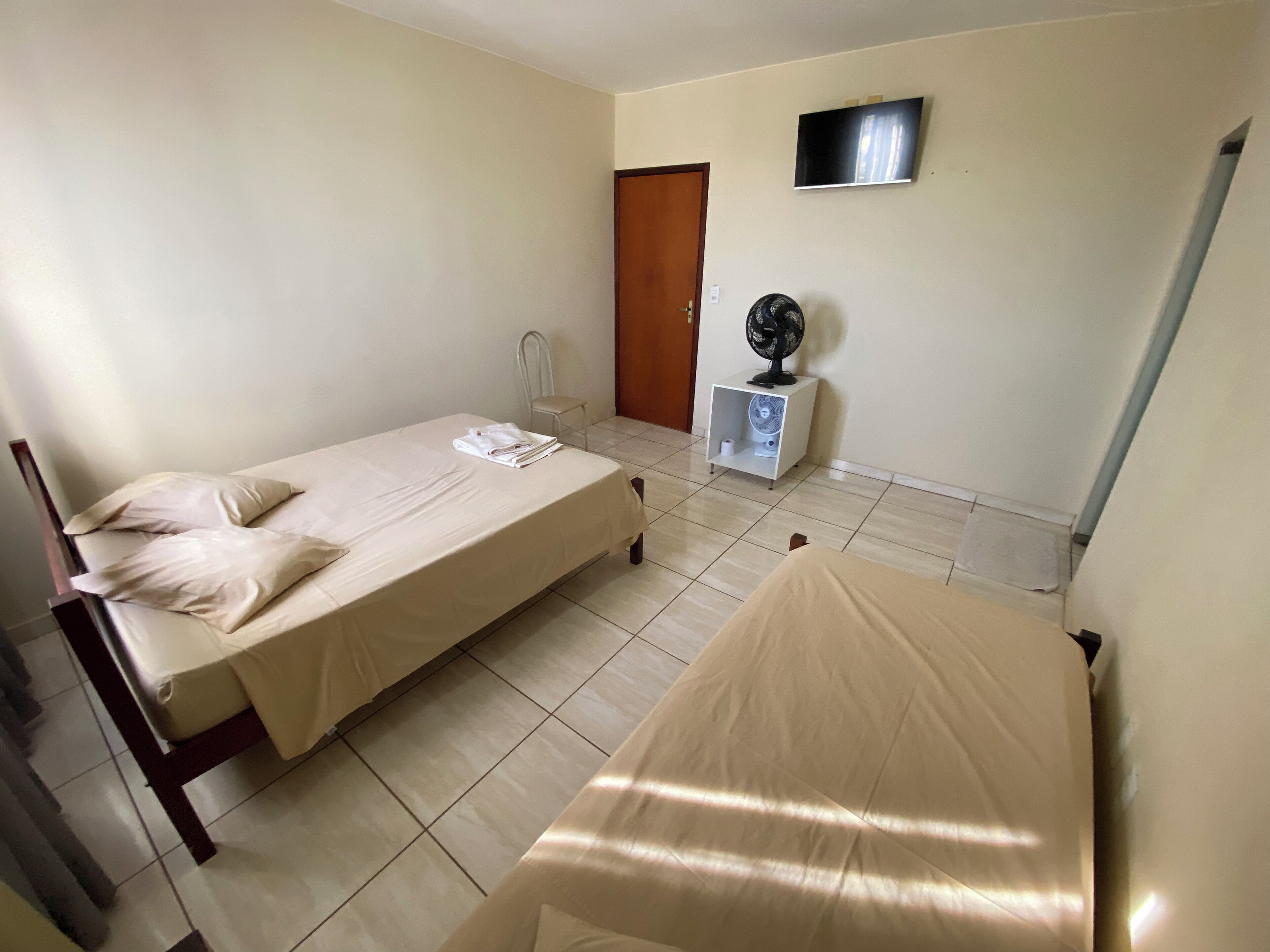 Une chambre avec deux lits simples, une chaise, une petite table avec un ventilateur et un téléviseur fixé au mur.