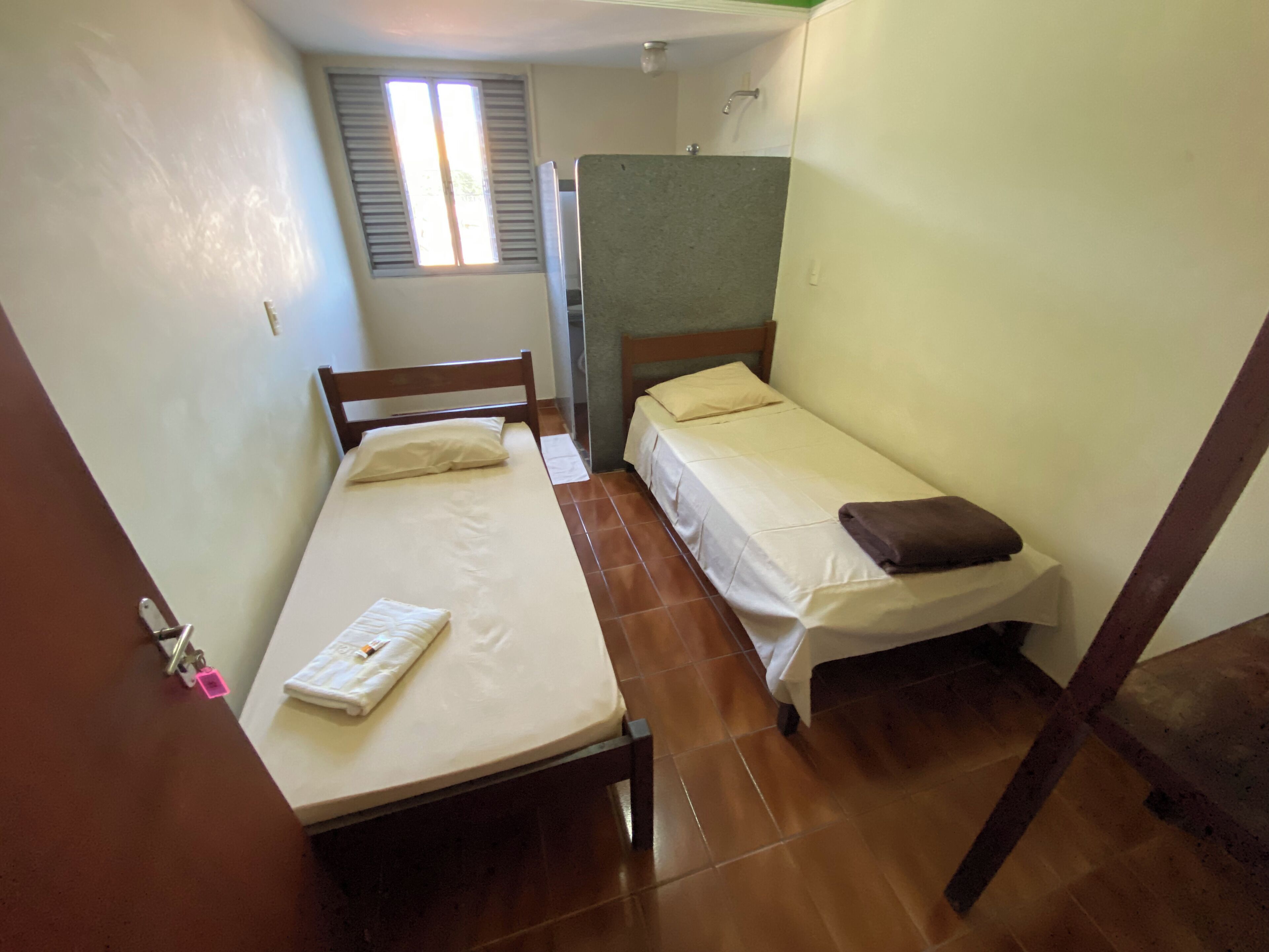 Une petite chambre compacte avec deux lits simples, une fenêtre avec volets, un espace douche et un sol carrelé.