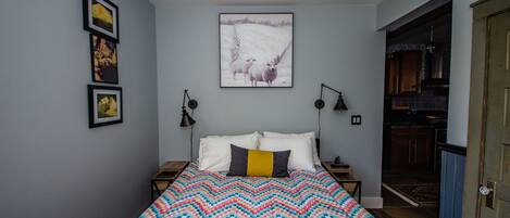 A bedroom with a bed, a ceiling fan, a wall-mounted picture, and a door leading to a kitchen.