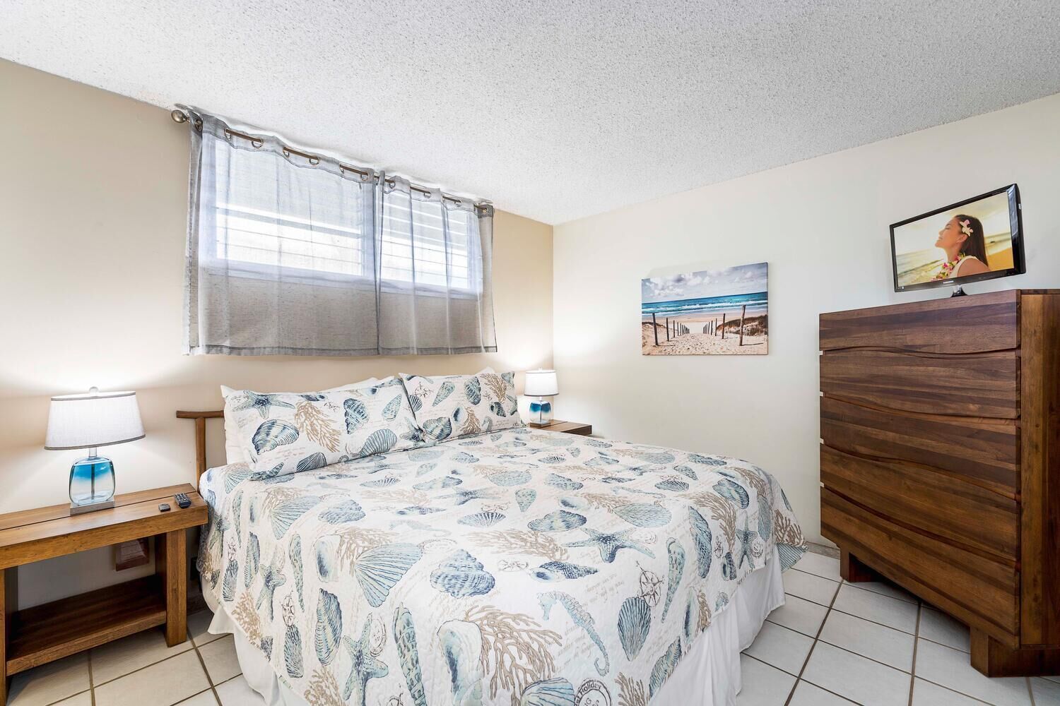 A bedroom with a bed, a wooden dresser, a nightstand, a lamp, and a beach-themed painting on the wall.