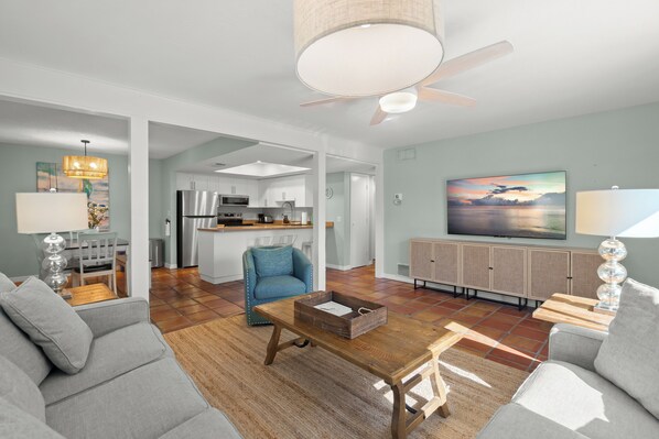 Ein geräumiger Wohnbereich mit Couchtisch aus Holz, grauen Sofas, einem blauen Sessel und einer Küche im Hintergrund.
