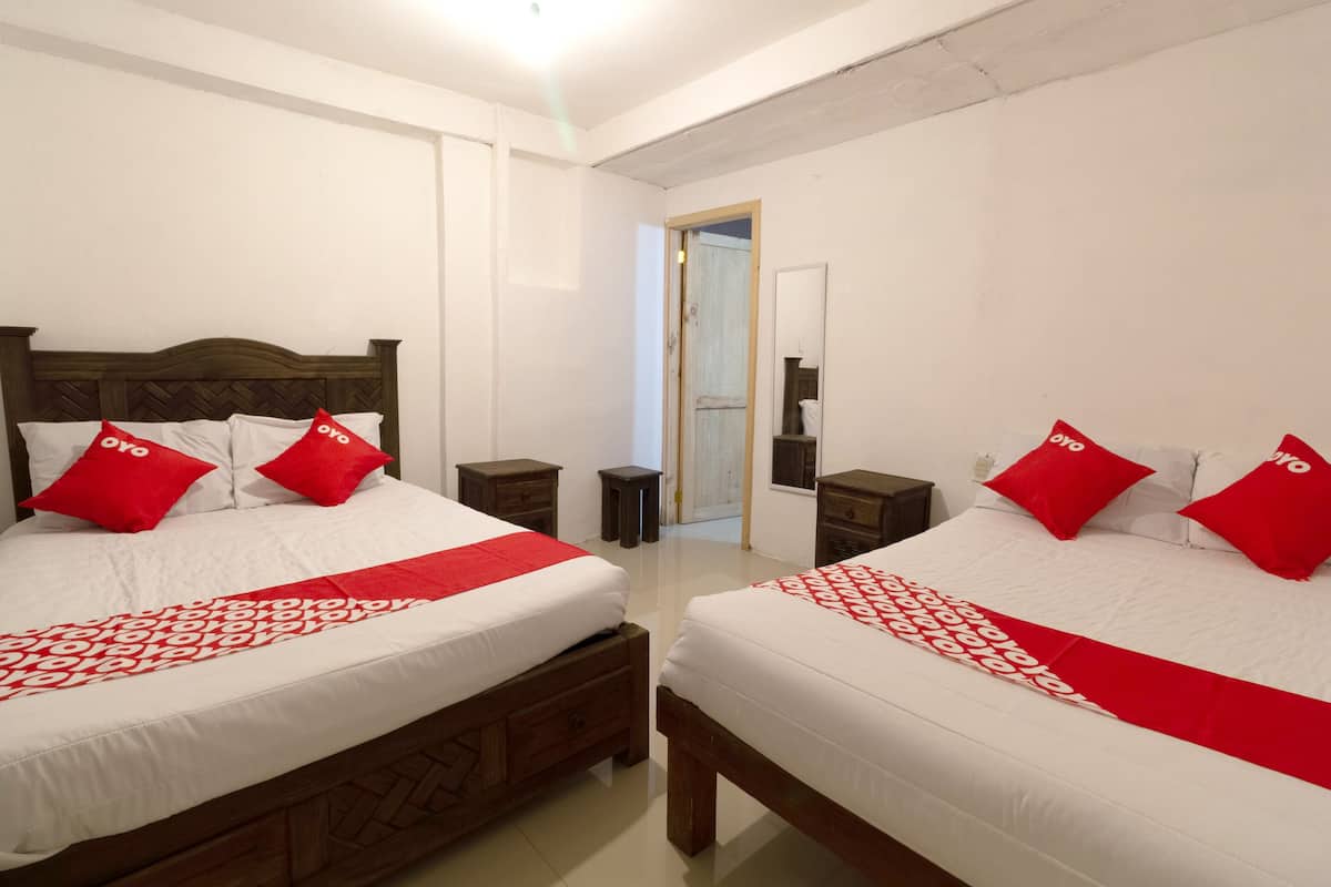 Habitación de hotel con dos camas, cada una con cabecero de madera, ropa de cama blanca y almohadas rojas. Hay mesitas de noche, un taburete pequeño y una puerta que da a otra habitación.