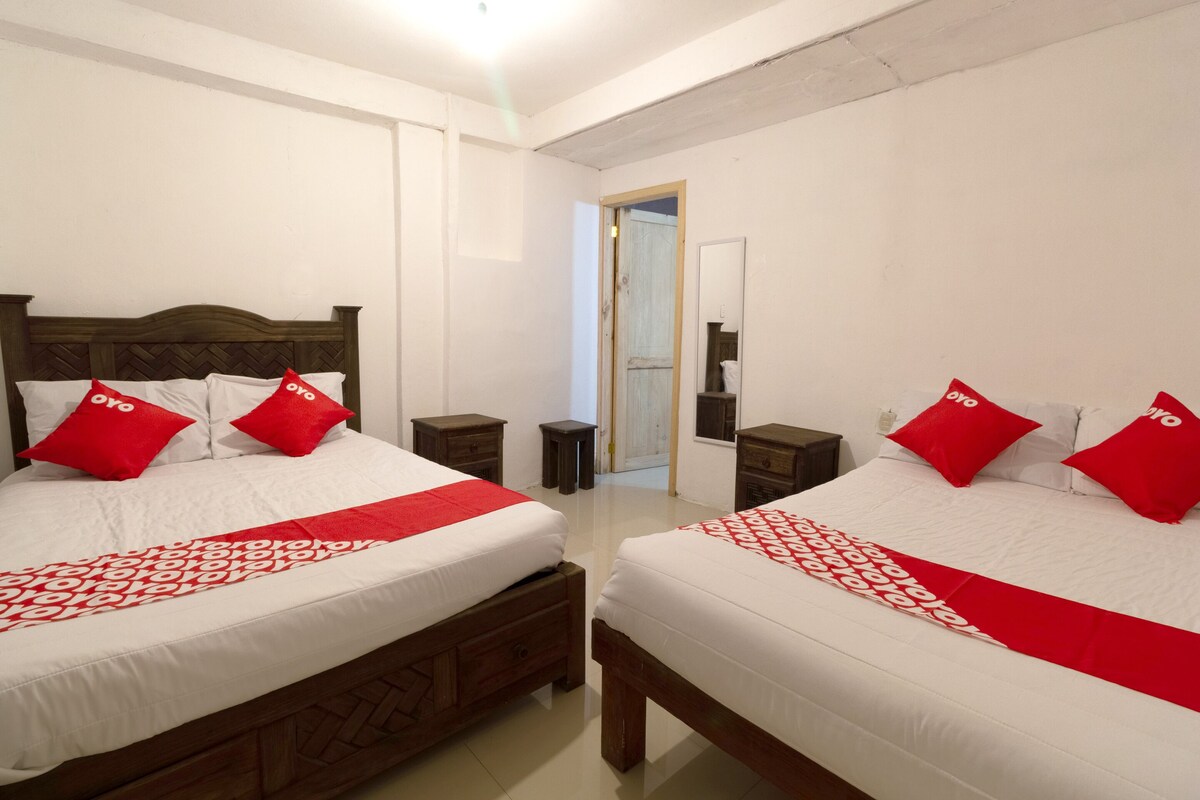 Habitación de hotel con dos camas, cada una con cabecero de madera, ropa de cama blanca y almohadas rojas. Hay mesitas de noche, un taburete pequeño y una puerta que da a otra habitación.