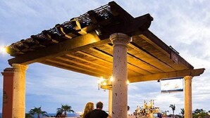 Un área de comedor al aire libre con una pérgola, mesas y sillas, rodeada de columnas.