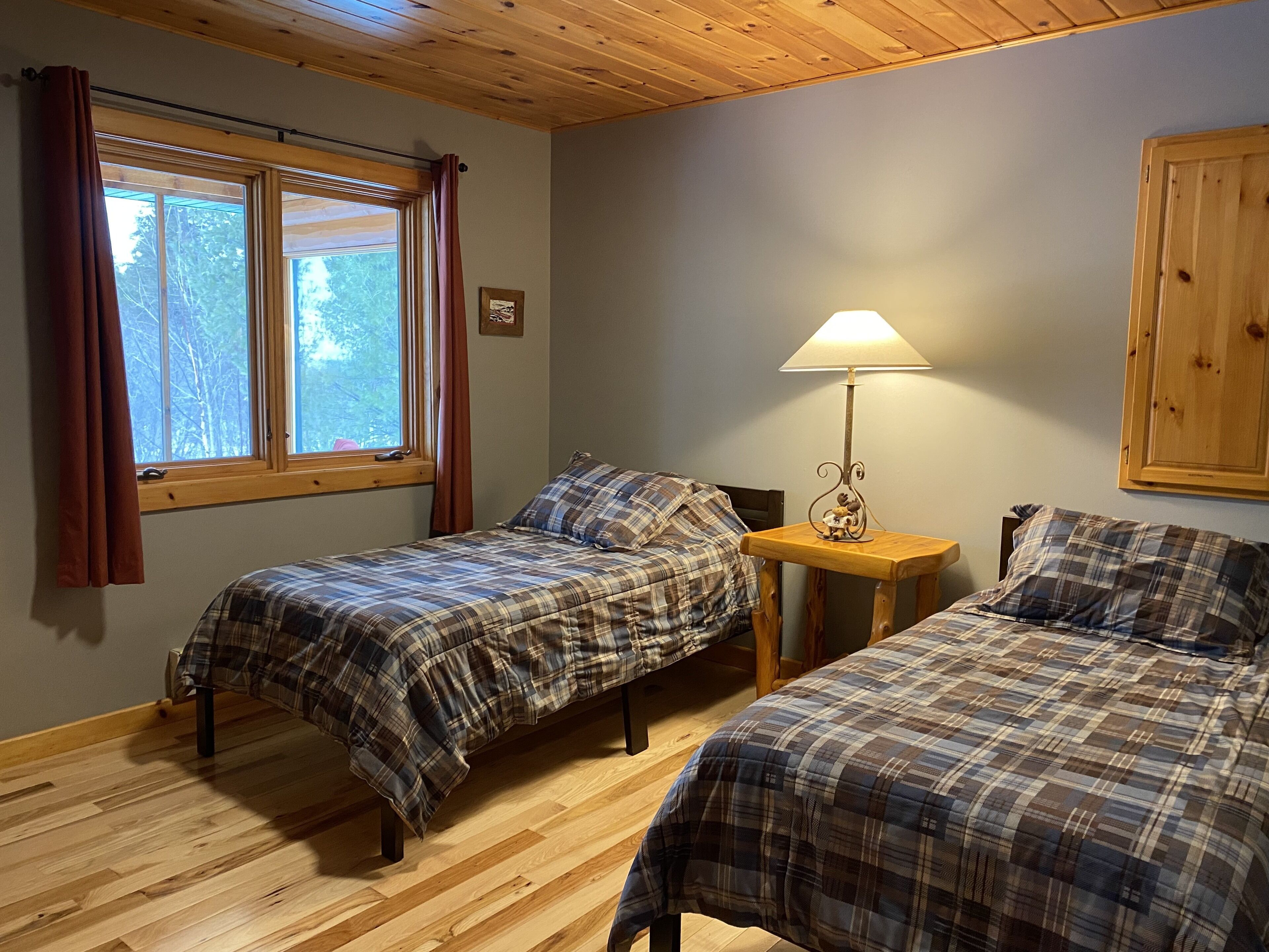 Une chambre avec deux lits, un plancher en bois, une fenêtre avec des rideaux, un meuble fixé au mur et une table avec une lampe.