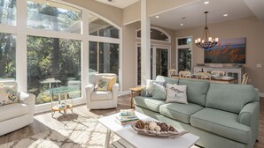 A spacious living room with large windows, a green sofa, white armchairs, and a coffee table with decorative items.
