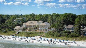 A beachfront property with a large house, a sandy beach with umbrellas, and dense greenery in the background.